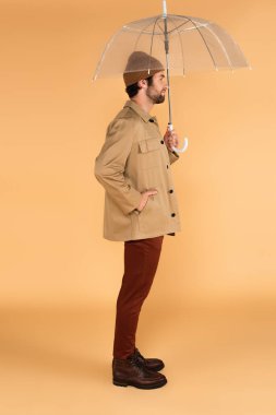 side view of man with hand in pocket of stylish jacket standing under umbrella on beige