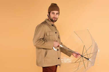 man in stylish autumn outfit looking at camera while folding umbrella isolated on beige