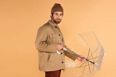 man in stylish autumn outfit looking at camera while folding umbrella isolated on beige clipart