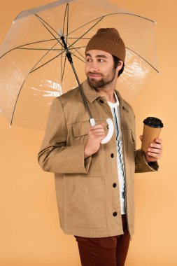 positive man with paper cup looking back under transparent umbrella isolated on beige