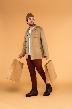 full length view of man in trendy autumn clothes walking with shopping bags on beige