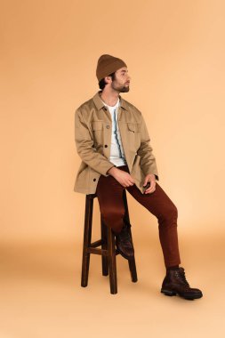 stylish man in jacket, brown pants and beanie looking away on high stool on beige