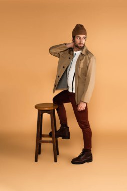 young man in fashionable clothes looking away while posing near wooden stool on beige