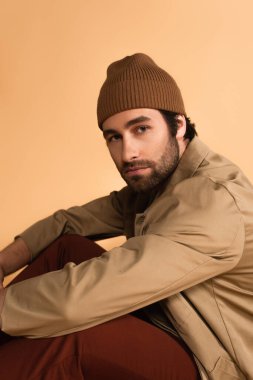 young man in brown beanie and jacket looking at camera isolated on beige