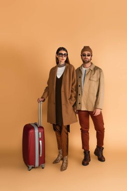 young fashionable couple in sunglasses near suitcase on beige background