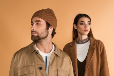 trendy man in beanie looking aside near blurred stylish woman isolated on beige