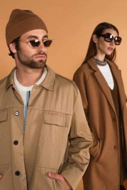 man in stylish sunglasses standing with hand in pocket of jacket near blurred woman isolated on beige