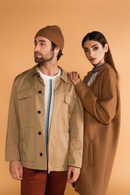 fashionable woman leaning on shoulder of trendy man in beanie isolated on beige