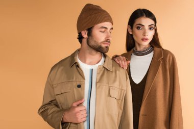 stylish woman touching shoulder of trendy man in beanie isolated on beige