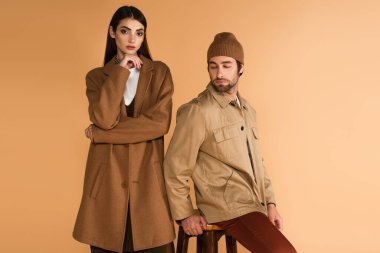 woman in brown coat standing near stylish man sitting on stool isolated on beige