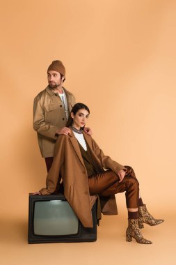man in beanie and jacket touching shoulders of trendy woman sitting on vintage tv set on beige background