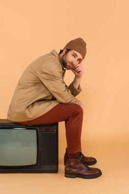 young man in fashionable autumn clothes sitting on vintage tv set on beige