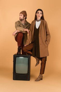 woman in stylish coat near man in jacket and beanie stepping on vintage tv set on beige background clipart