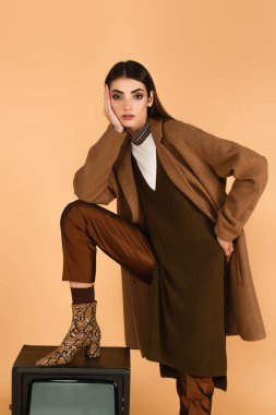 fashionable woman stepping on retro tv set while posing with hand on hip isolated on beige