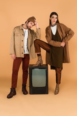 woman in trendy clothes stepping on vintage tv set near man touching beanie on beige background