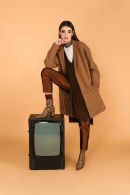 trendy woman with hand in pocket of trendy coat stepping on vintage tv on beige 