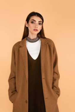 pretty young woman holding hands in pockets of brown coat isolated on beige