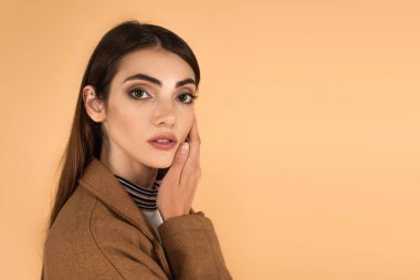young brunette woman touching face while looking at camera isolated on beige