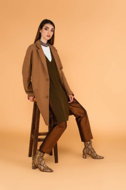 pretty brunette woman in fashionable autumn outfit sitting on wooden stool on beige 