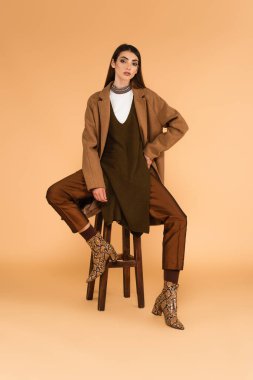 young woman in stylish coat and boots sitting on wooden stool on beige background