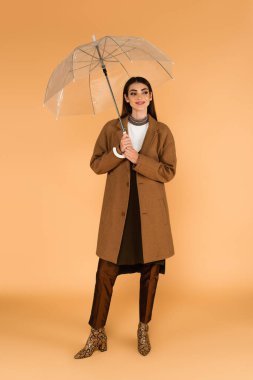 stylish woman smiling while looking away under transparent umbrella on beige 