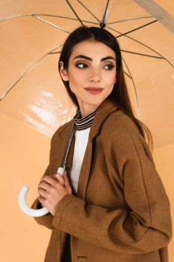 positive young woman in brown coat looking away under umbrella isolated on beige