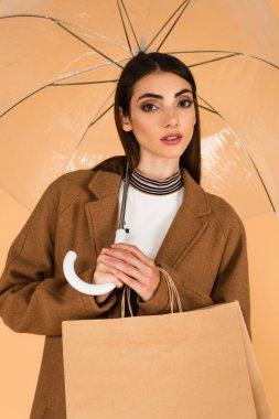 stylish woman looking at camera while standing under umbrella with shopping bags isolated on beige