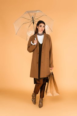 fashionable woman standing with shopping bags under transparent umbrella on beige 