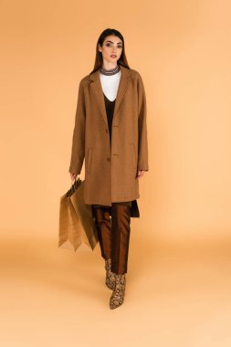 full length view of woman in trendy autumn clothes holding shopping bags on beige background