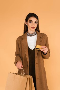 brunette woman in trendy coat holding credit card and shopping bags isolated on beige