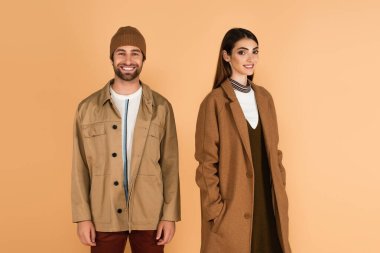 happy couple in trendy autumn clothes looking at camera isolated on beige clipart