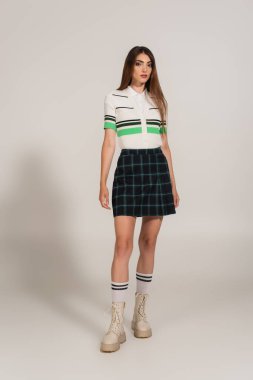 full length view of young woman in plaid skirt and high boots on grey background