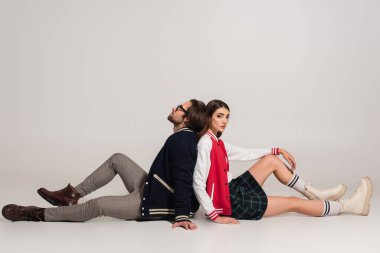 young stylish woman looking at camera while sitting back to back with trendy man on grey 