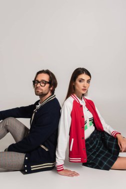 young couple in trendy jackets looking at camera while sitting on grey background