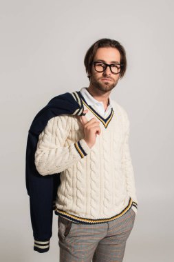 trendy man in white jumper and eyeglasses posing with hand in pocket isolated on grey