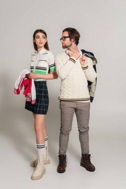man in eyeglasses and white pullover looking at trendy young woman on grey background