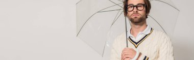 young man in white sweater and eyeglasses standing under transparent umbrella isolated on grey, banner