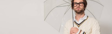 young man in white sweater and eyeglasses standing under transparent umbrella isolated on grey, banner clipart