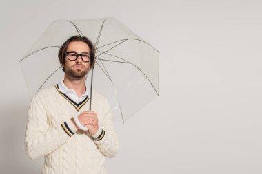 man in white knitted pullover standing under transparent umbrella isolated on grey