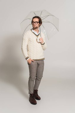 full length view of stylish man standing under umbrella with hand in pocket on grey 