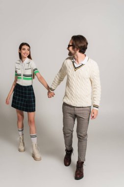happy and fashionable couple holding hands while walking on grey background