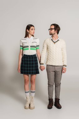 full length view of couple in stylish clothes holding hands and looking at each other on grey background