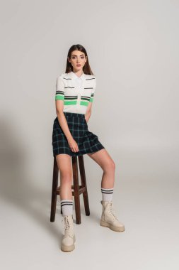 young woman in trendy clothes sitting on high wooden stool on grey background