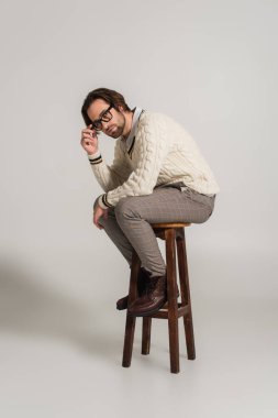 man in white jumper and trousers adjusting eyeglasses while sitting on grey background