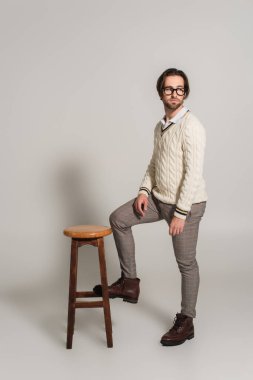 fashionable man in sweater and pants looking away near high, wooden stool on grey 