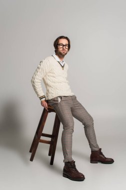 young man in white sweater and pants sitting on high wooden stool on grey background