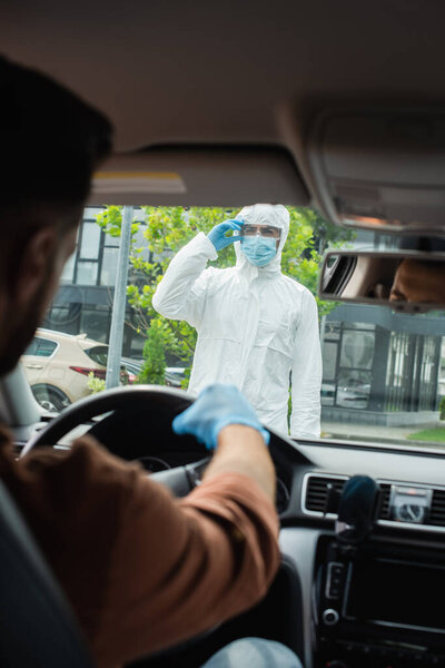 Medical worker in hazmat suit and goggles standing near blurred driver in car 