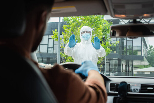 Medical worker in protective suit showing stop at blurred driver in auto 