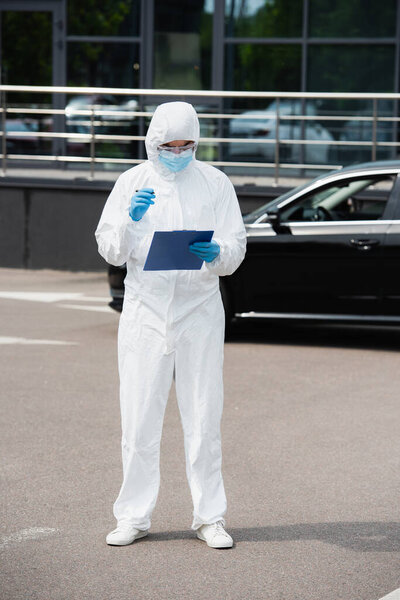 Medical worker in hazmat suit holding clipboard near blurred car outdoors 