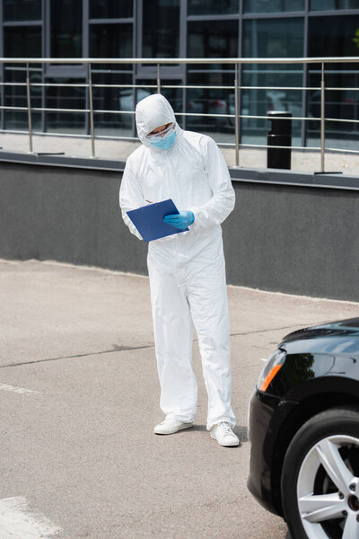 Border guard in medical mask and protective suit writing on clipboard and looking at car outdoors 